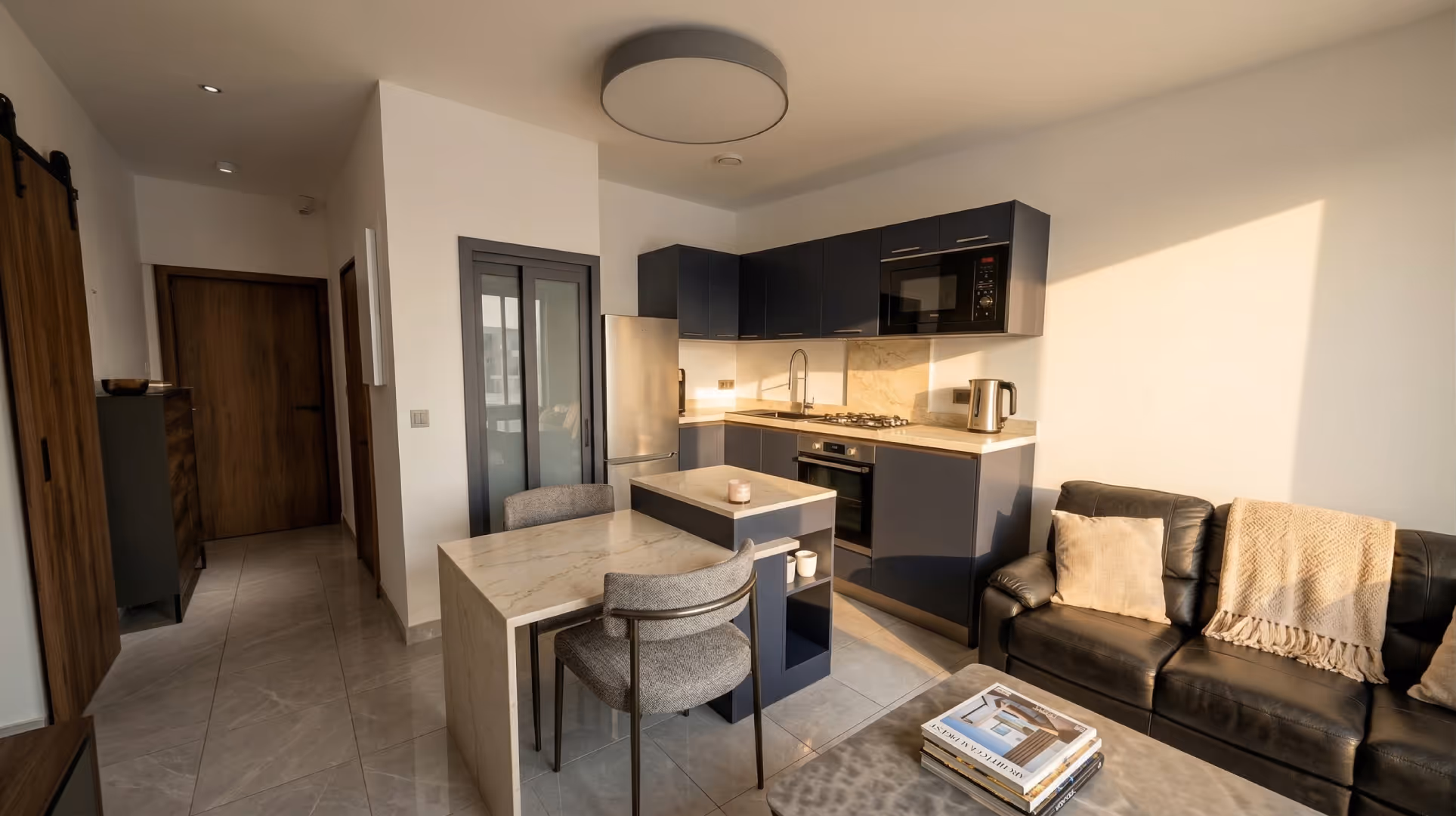 Cuisine moderne avec meubles sombres, réfrigérateur en acier inoxydable, table en marbre avec deux chaises, canapé en cuir noir avec coussins et plaid beige.